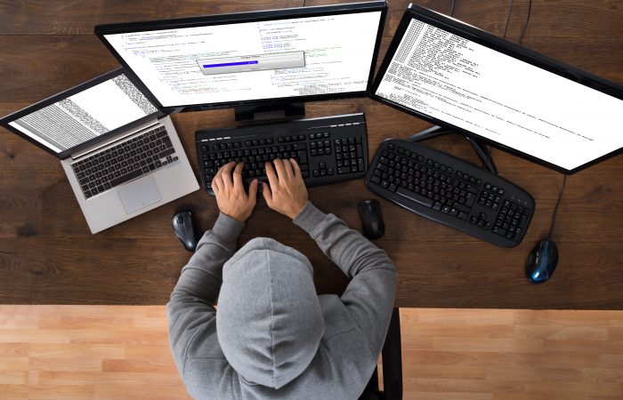 A hooded man at three computers hacking into databases