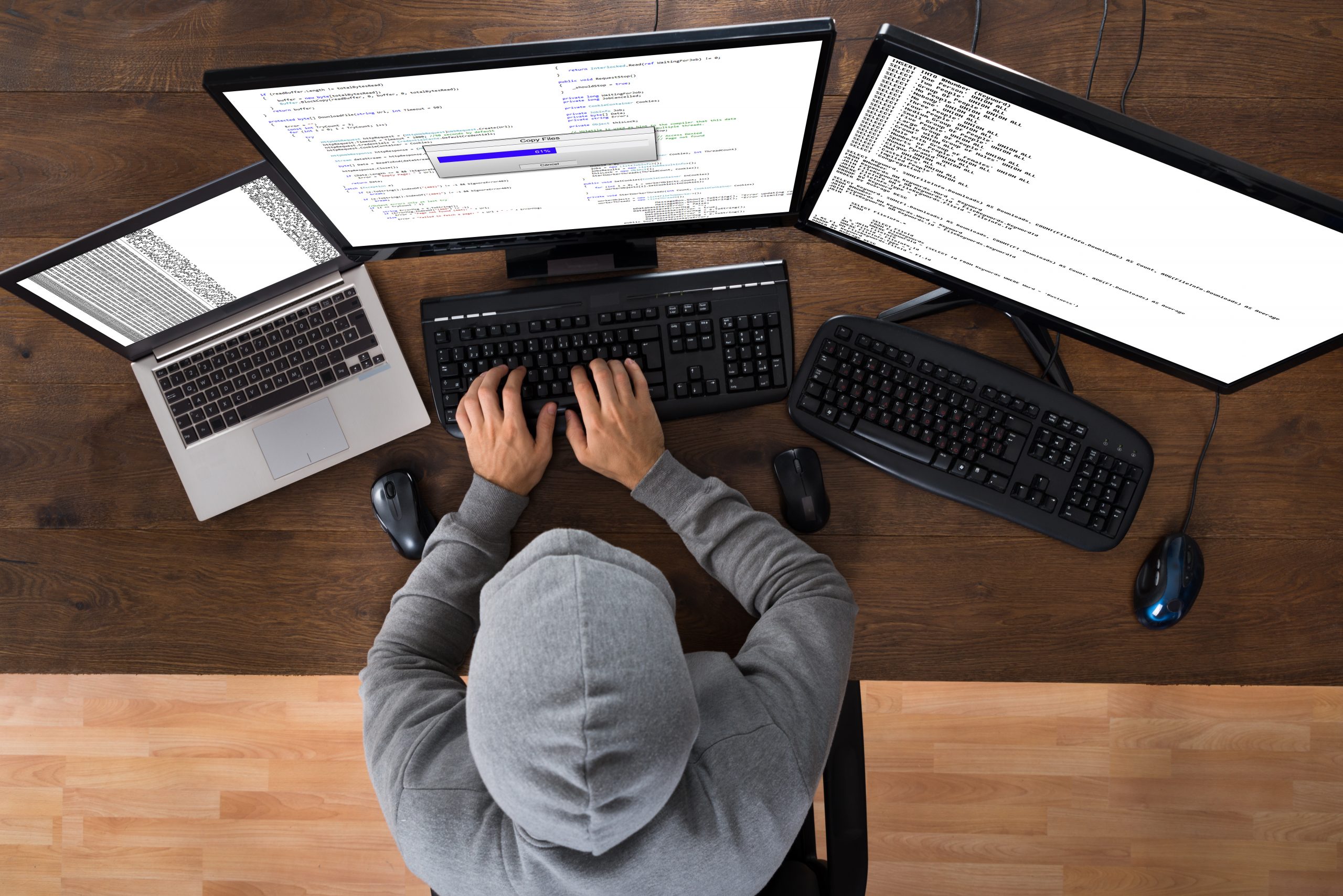 A hooded man at three computers hacking into databases