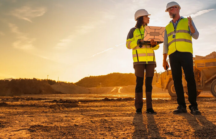 Two people in construction dress looking at laptop in front of an open cleared landscape