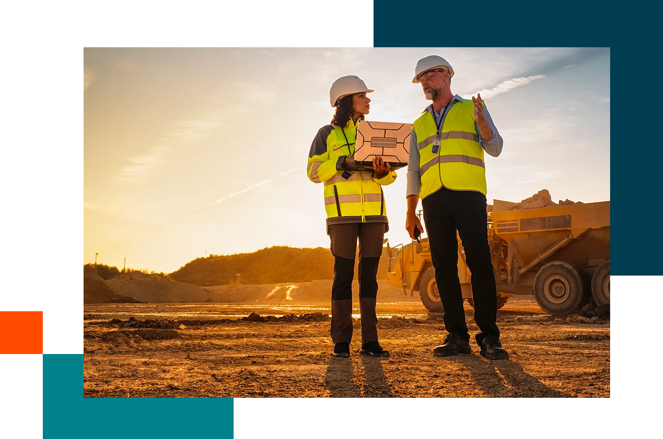 Two people in construction dress looking at laptop in front of an open cleared landscape