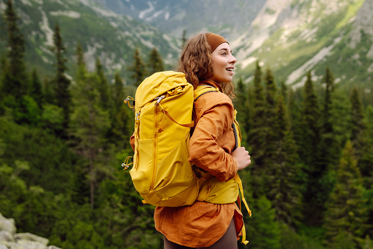 Woman with orange jacket and yellow backpack walking through a forest