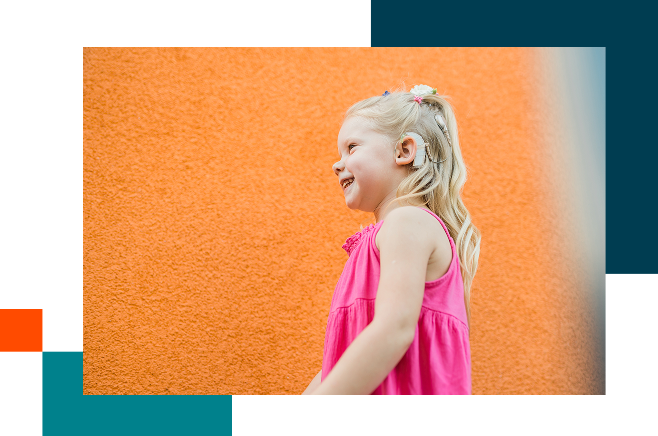 A young child with long blonde hair, wearing a pink sleeveless dress, standing in profile against an orange textured wall