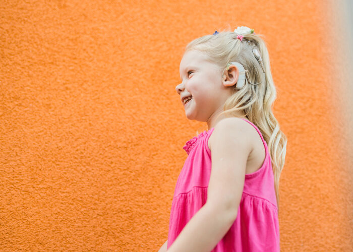 A young child with long blonde hair, wearing a pink sleeveless dress, standing in profile against an orange textured wall