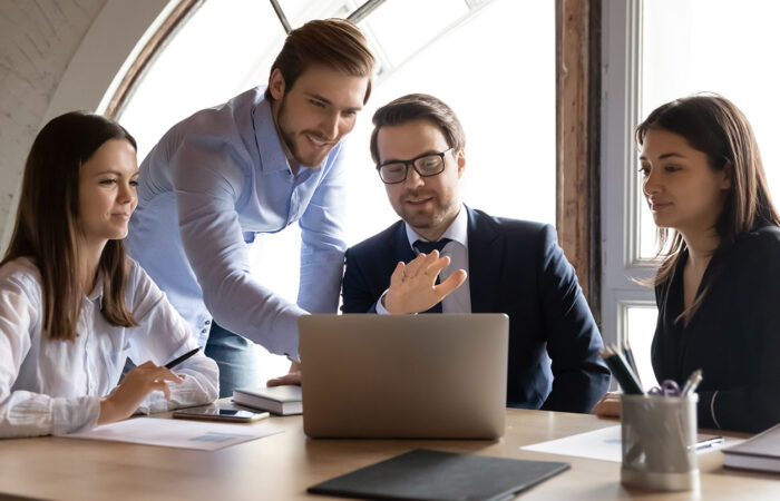 Four people discussing around a table in an office, with one pointing at a laptop.