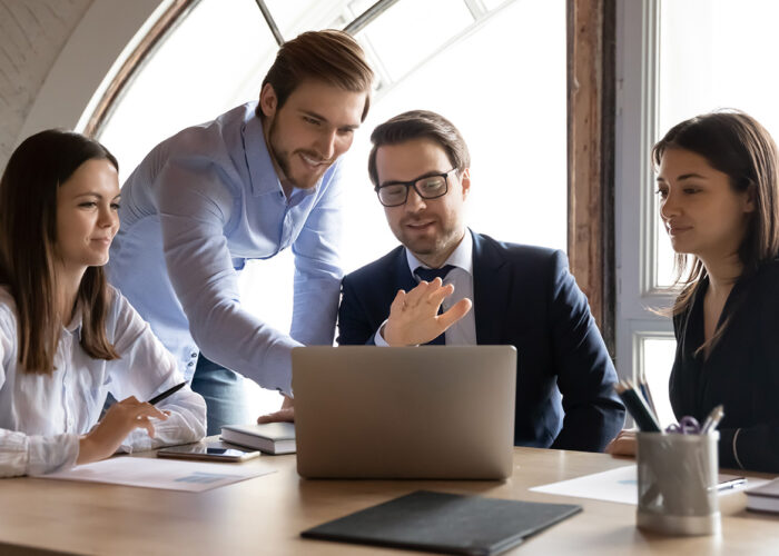 Four people discussing around a table in an office, with one pointing at a laptop.