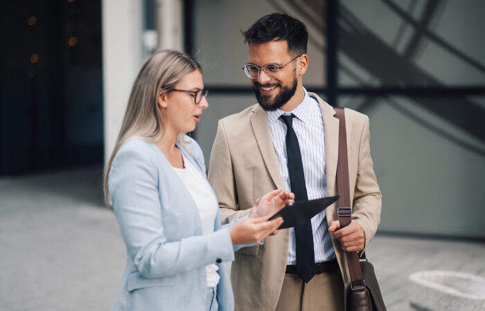 Two people in business attire stand outside a building, with one showing something on a tablet to the other; faces are blurred.