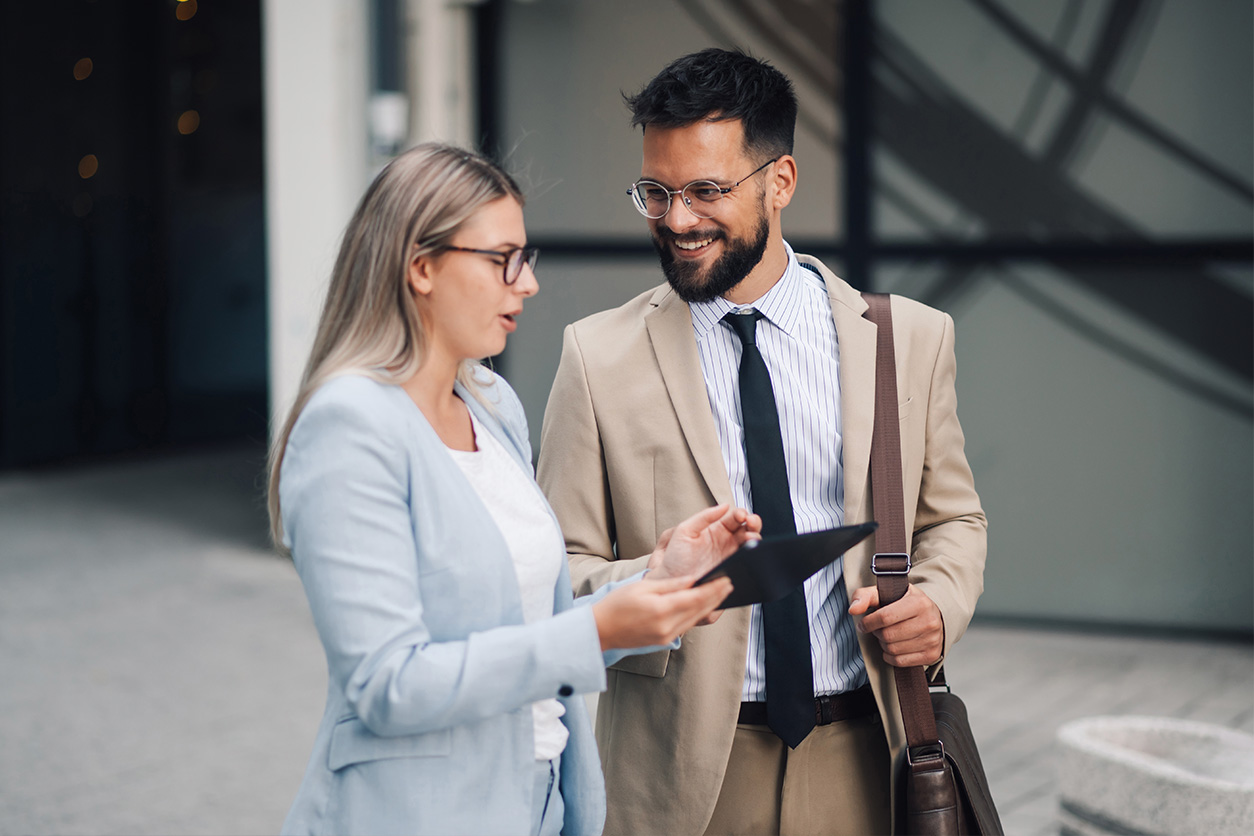 Two people in business attire stand outside a building, with one showing something on a tablet to the other; faces are blurred.