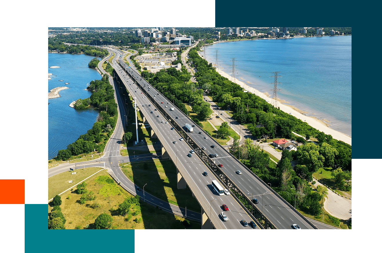 A multi-lane highway runs alongside a beach and forested area, with a cityscape visible in the distance under a partly cloudy sky.