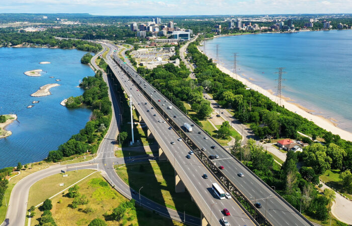 A multi-lane highway runs alongside a beach and forested area, with a cityscape visible in the distance under a partly cloudy sky.