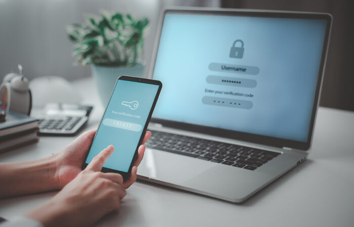 A person holds a smartphone showing a verification code while logging in on a laptop using two-factor authentication.