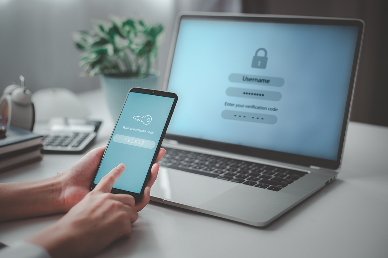 A person holds a smartphone showing a verification code while logging in on a laptop using two-factor authentication.