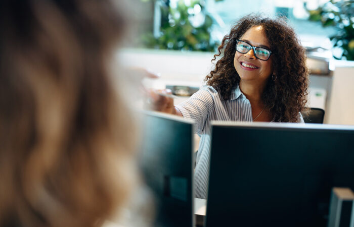 Two people seated at desks in an office, working on computers; one person's face is pixelated for privacy.