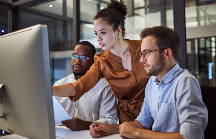 Three people in a modern office setting gathered around a computer monitor. One person stands and points at the screen while holding a paper, and two others sit attentively, suggesting a collaborative discussion or analysis.