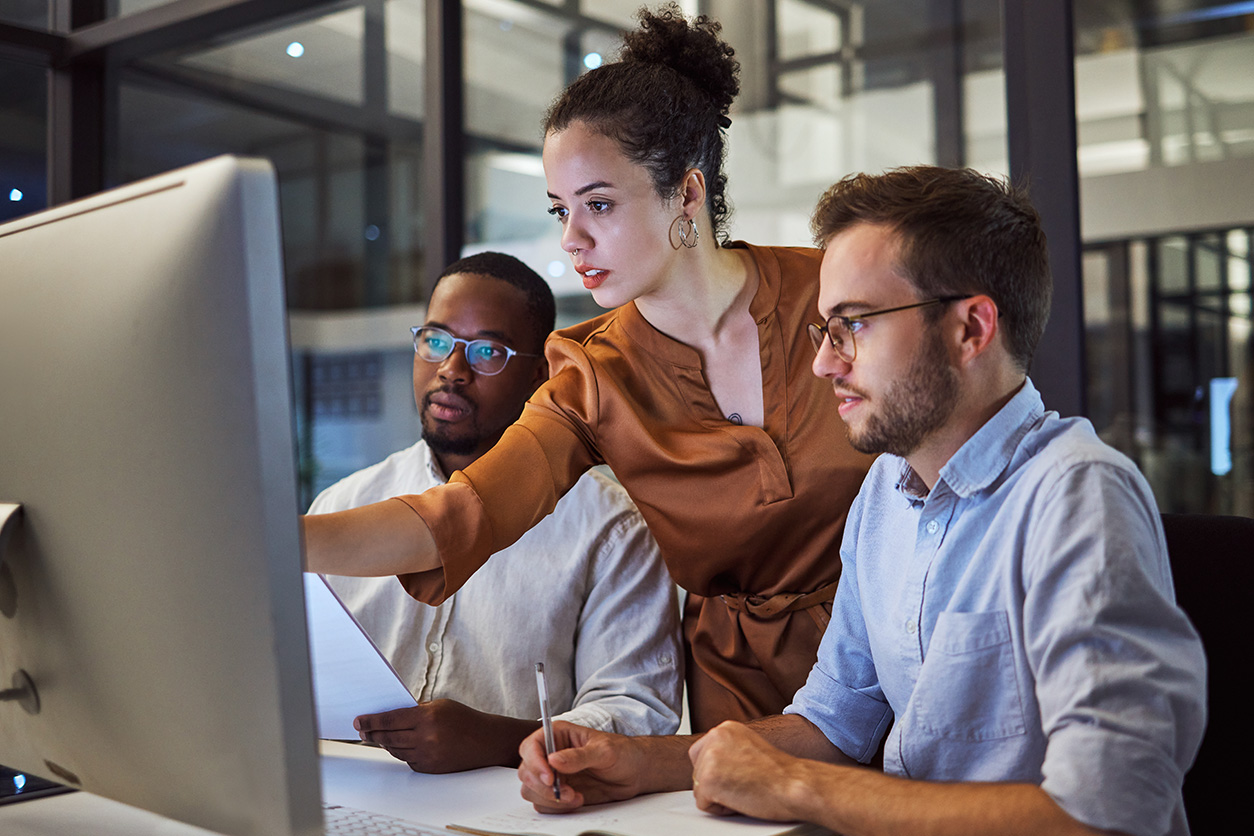 Three people in a modern office setting gathered around a computer monitor. One person stands and points at the screen while holding a paper, and two others sit attentively, suggesting a collaborative discussion or analysis.