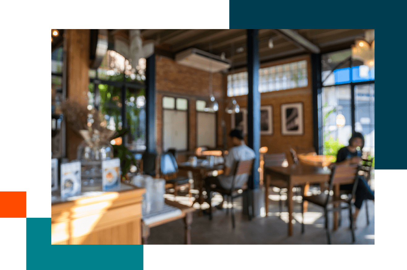 Interior of a cozy café with wooden tables and chairs, warm lighting, and large windows letting in natural light. Menus and decorative items are visible on the counter in the foreground, with framed artwork on the brick wall in the background.