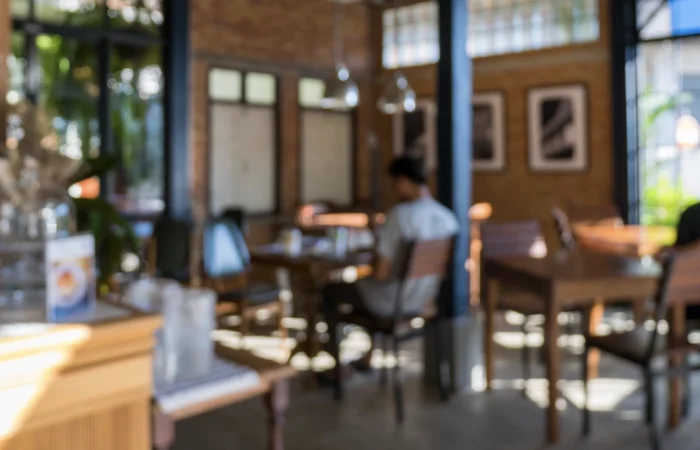 Interior of a cozy café with wooden tables and chairs, warm lighting, and large windows letting in natural light. Menus and decorative items are visible on the counter in the foreground, with framed artwork on the brick wall in the background.