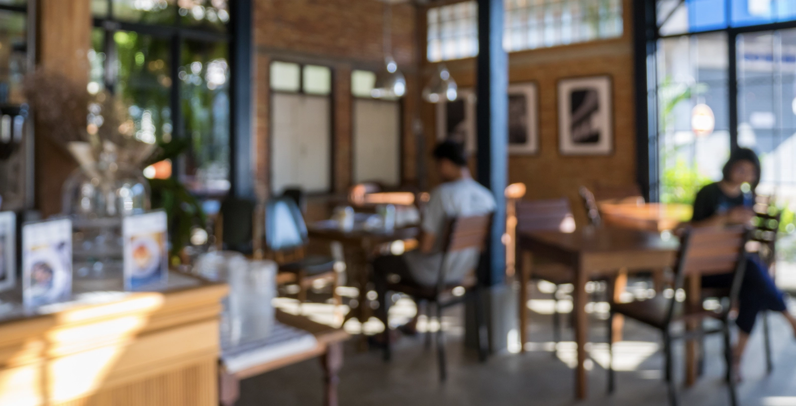 Interior of a cozy café with wooden tables and chairs, warm lighting, and large windows letting in natural light. Menus and decorative items are visible on the counter in the foreground, with framed artwork on the brick wall in the background.