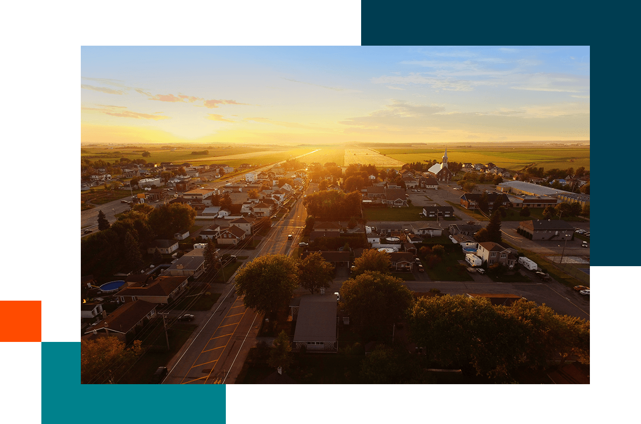 A small rural town at sunset, with rows of houses, tree-lined streets, and a church visible on the right. The golden sunlight stretches across open farmland in the background, creating a warm, glowing horizon