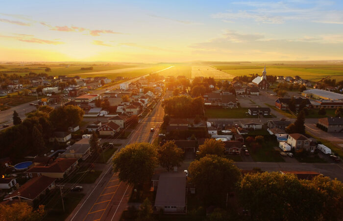 A small rural town at sunset, with rows of houses, tree-lined streets, and a church visible on the right. The golden sunlight stretches across open farmland in the background, creating a warm, glowing horizon