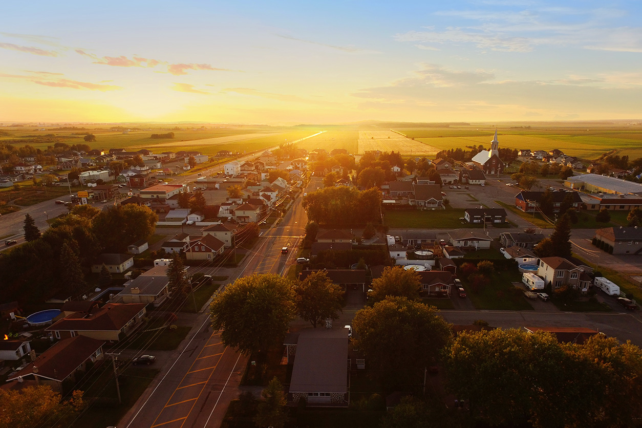A small rural town at sunset, with rows of houses, tree-lined streets, and a church visible on the right. The golden sunlight stretches across open farmland in the background, creating a warm, glowing horizon