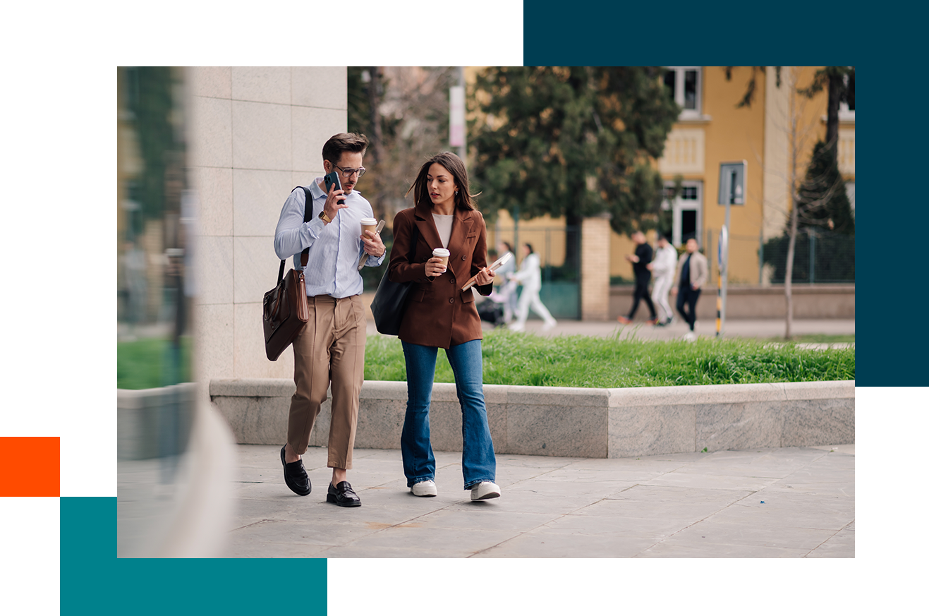 Two people walking outdoors on a paved area, holding drinks and talking, with buildings and greenery in the background.