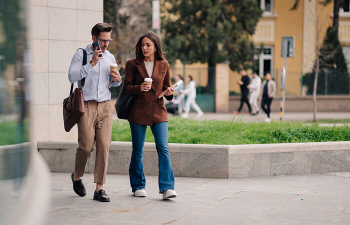 Two people walking outdoors on a paved area, holding drinks and talking, with buildings and greenery in the background.