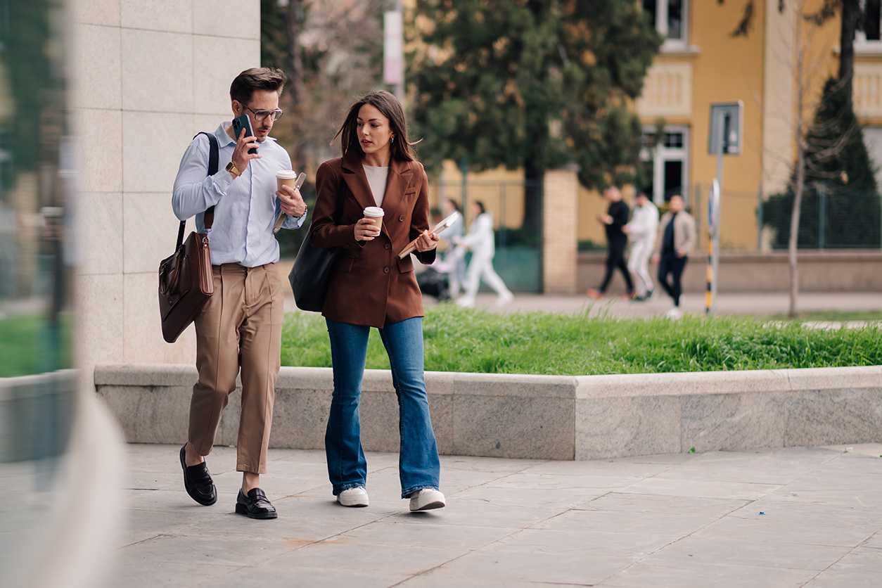 Two people walking outdoors on a paved area, holding drinks and talking, with buildings and greenery in the background.