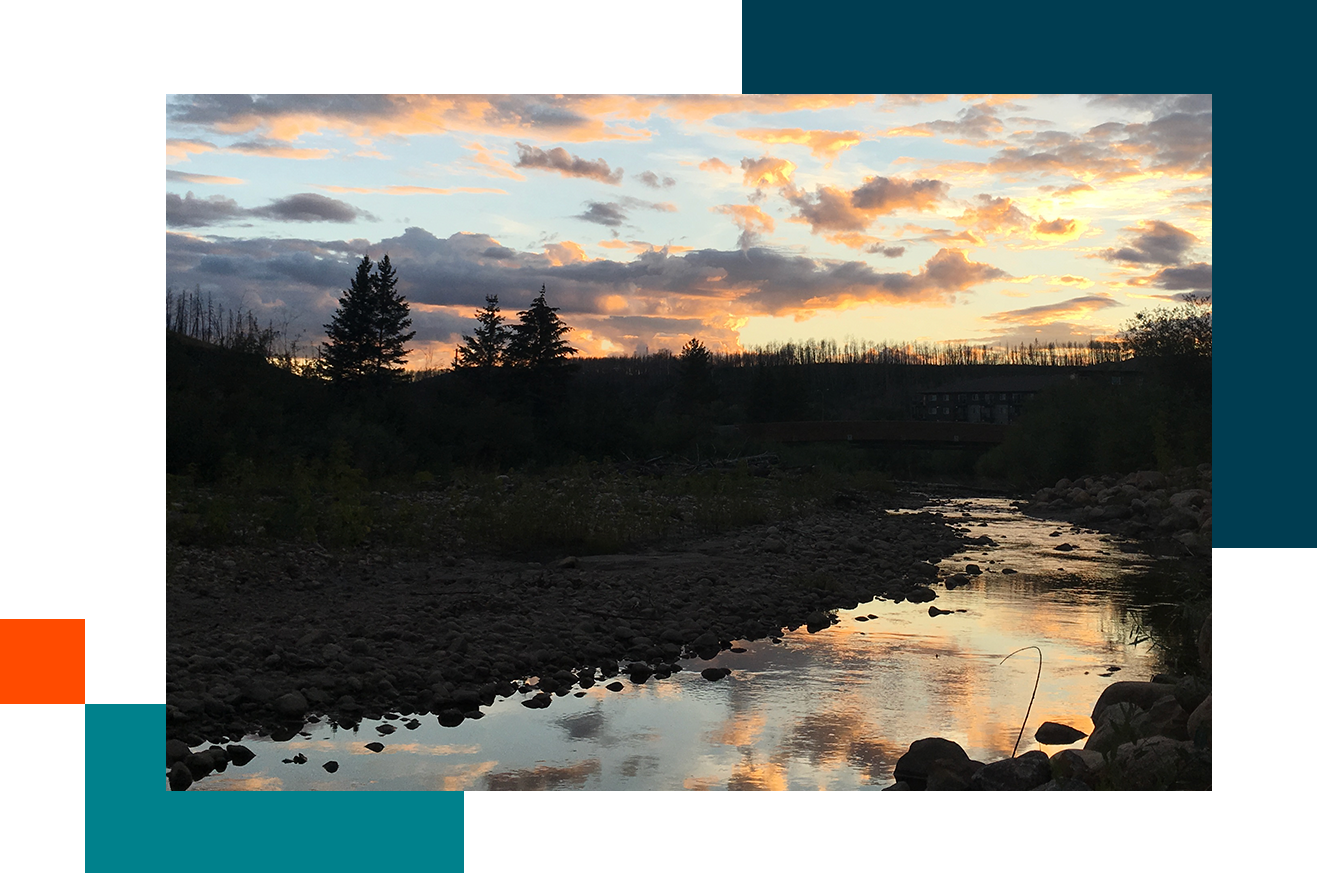Sunset over a rocky riverbed with trees silhouetted against a colorful sky of orange, pink, and blue clouds.