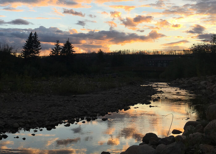 Sunset over a rocky riverbed with trees silhouetted against a colorful sky of orange, pink, and blue clouds.