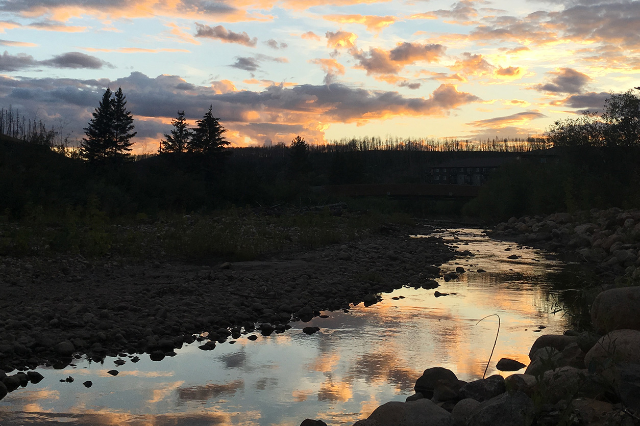 Sunset over a rocky riverbed with trees silhouetted against a colorful sky of orange, pink, and blue clouds.