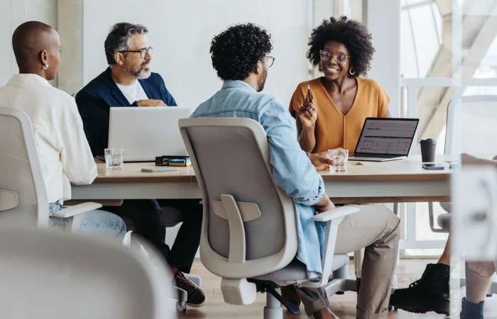 Group of people sitting around a conference table in a modern office, engaged in discussion with laptops and notebooks on the table.