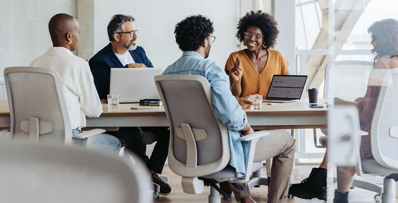 Group of people sitting around a conference table in a modern office, engaged in discussion with laptops and notebooks on the table.