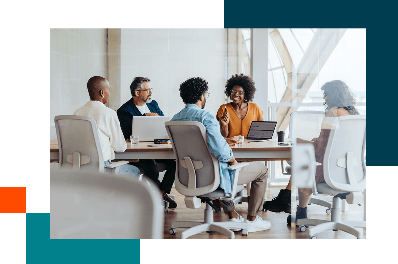 Group of people sitting around a conference table in a modern office, engaged in discussion with laptops and notebooks on the table.