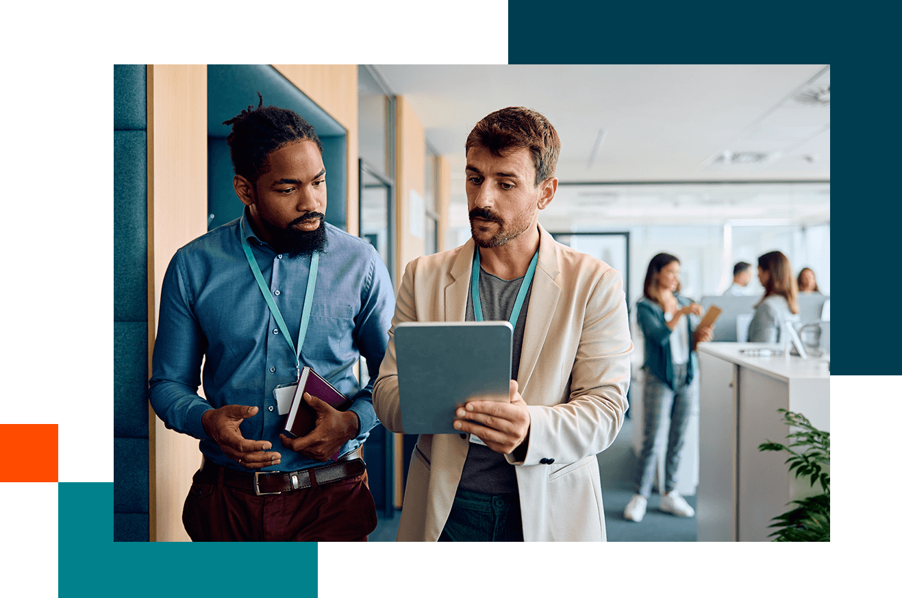 Two people standing in an office hallway reviewing information on a tablet while colleagues work in the background.