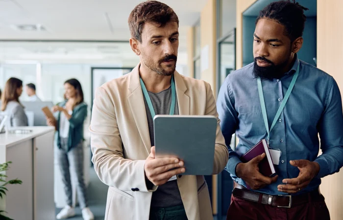 Two people standing in an office hallway reviewing information on a tablet while colleagues work in the background.