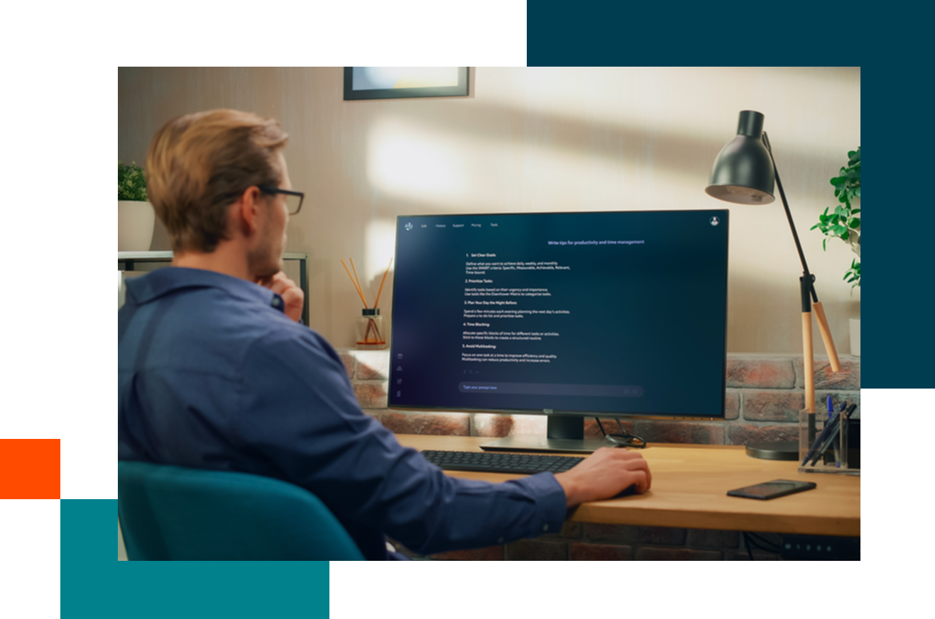 A person sits at a desk working on a large monitor displaying a dark-themed interface, with a keyboard, mouse, phone, and desk lamp nearby.