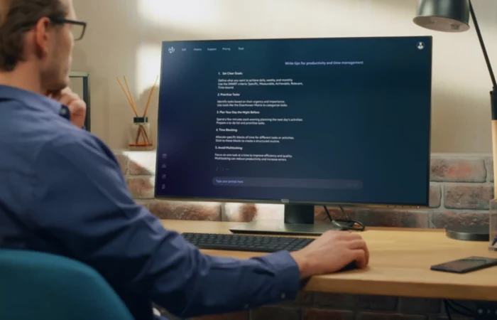 A person sits at a desk working on a large monitor displaying a dark-themed interface, with a keyboard, mouse, phone, and desk lamp nearby.