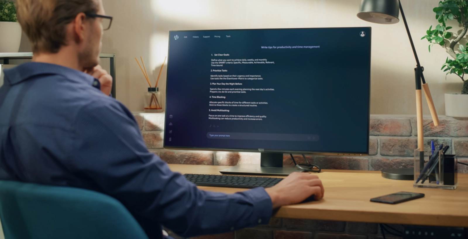 A person sits at a desk working on a large monitor displaying a dark-themed interface, with a keyboard, mouse, phone, and desk lamp nearby.