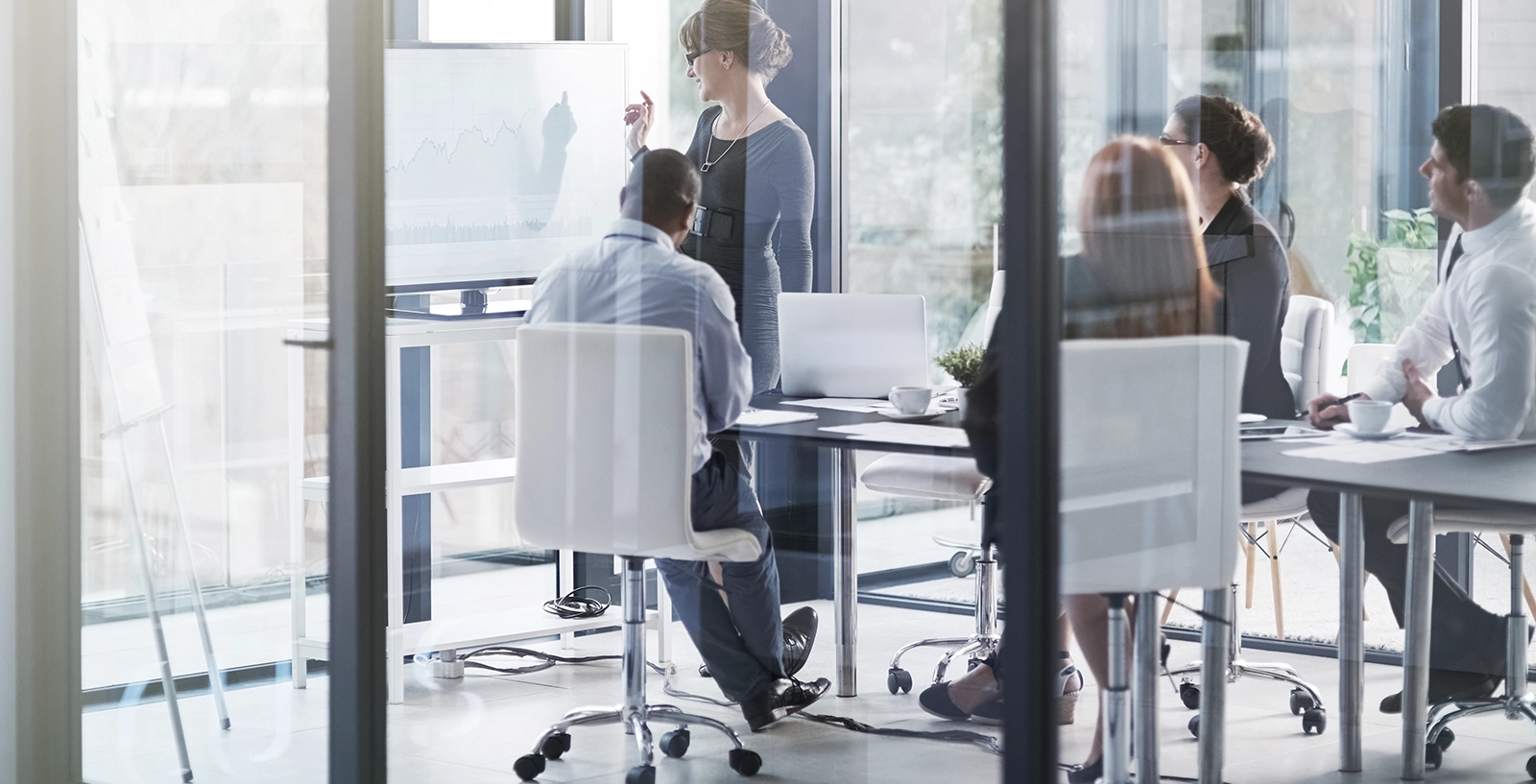Business meeting with six people and a presenter pointing at a graph on a screen.