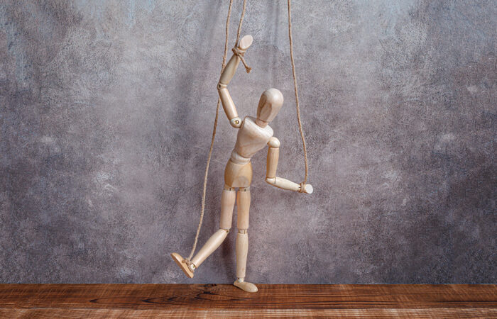 Wooden mannequin suspended by strings above a table, posed like a marionette against a textured gray background.