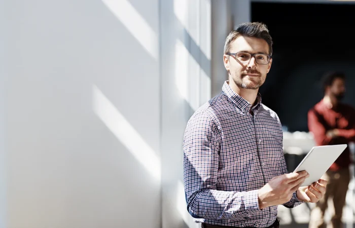Person standing indoors holding a tablet, with other people blurred in the background.