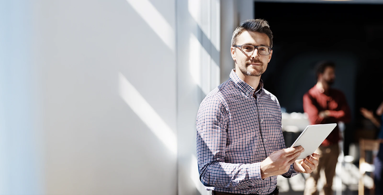 Person standing indoors holding a tablet, with other people blurred in the background.