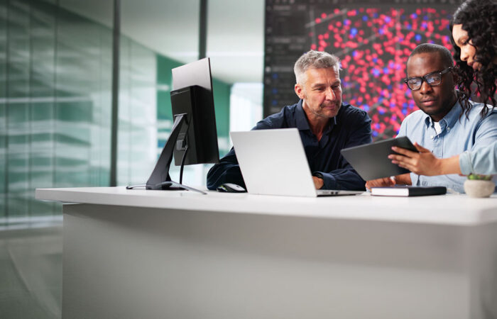 Professionals reviewing data on a tablet and laptop at a modern office desk with a large data visualization screen behind them.