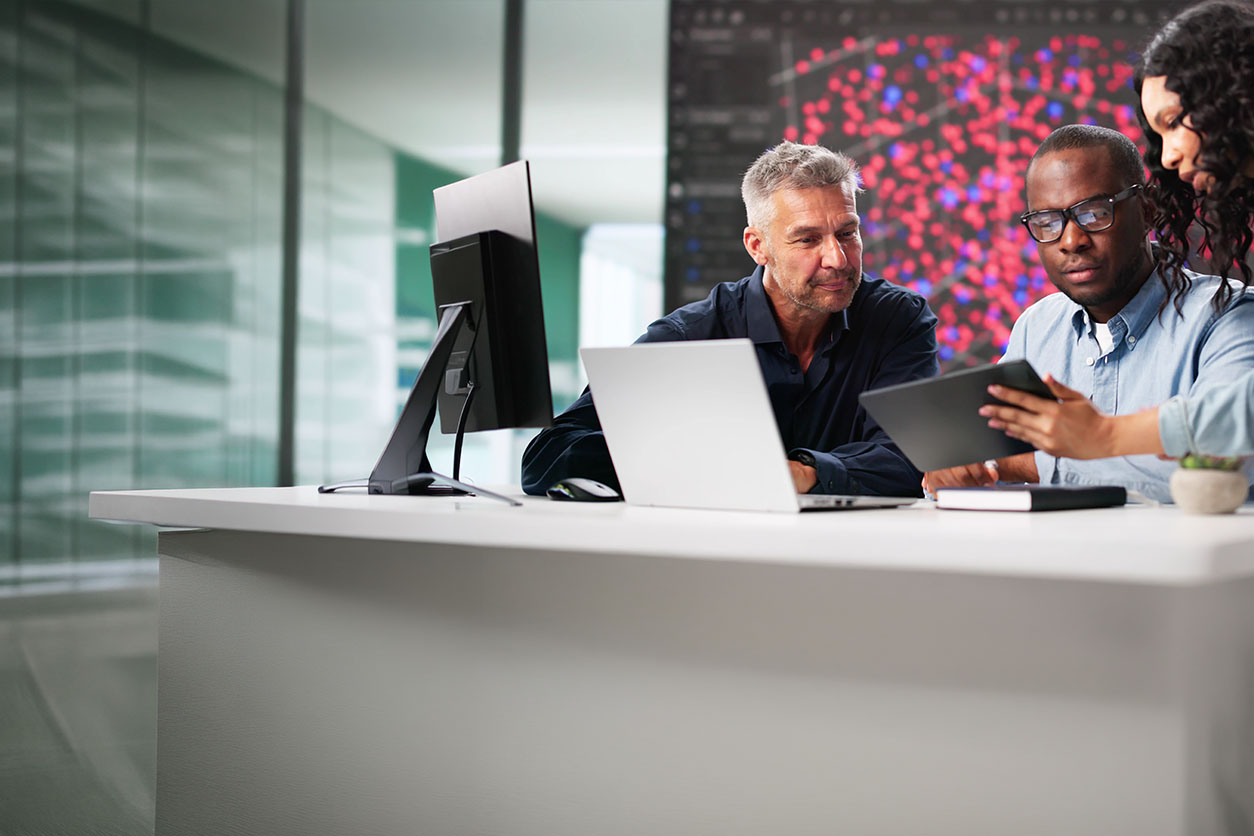 Professionals reviewing data on a tablet and laptop at a modern office desk with a large data visualization screen behind them.
