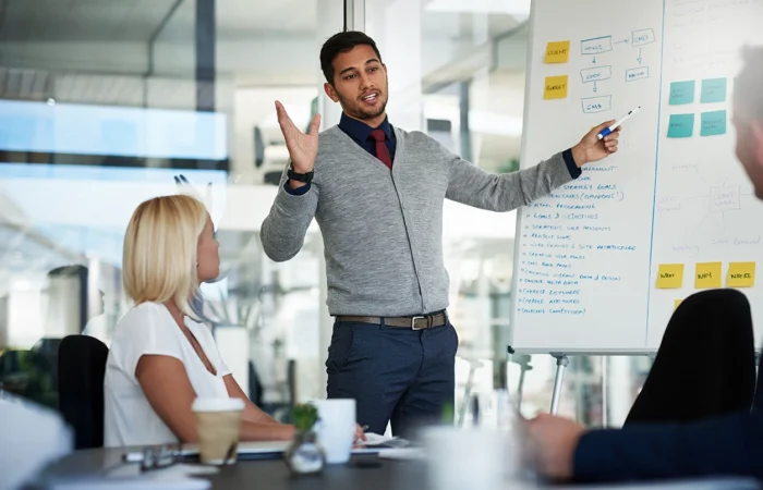 Person standing and pointing to a whiteboard while speaking to a small group in a meeting room.