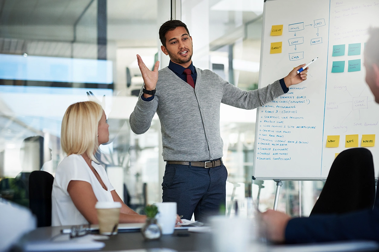Person standing and pointing to a whiteboard while speaking to a small group in a meeting room.