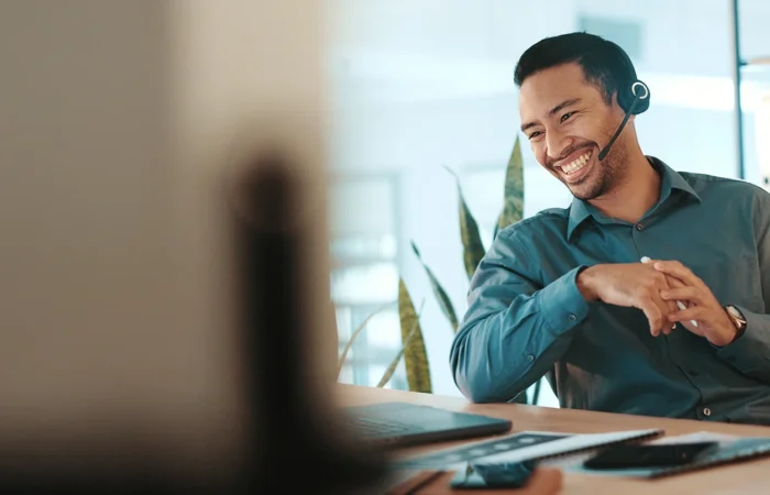 Person wearing a headset speaking at a desk with a laptop and smartphone, in a bright office setting with a plant in the background.