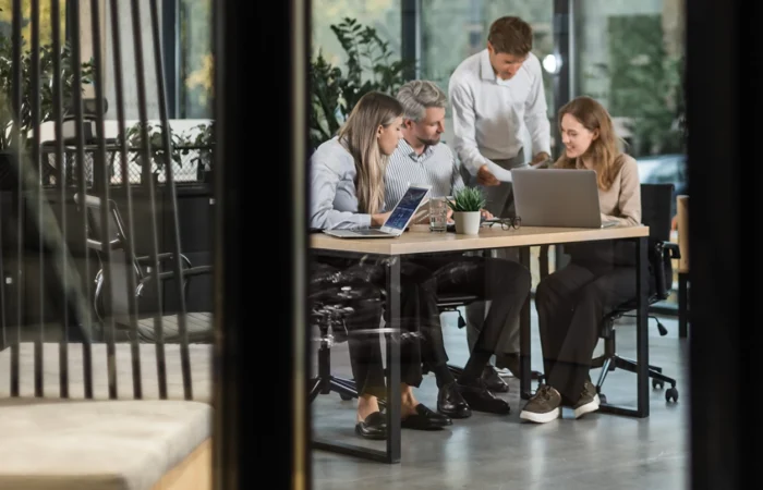 Four people collaborate around a table in a modern office meeting room, reviewing documents and working on laptops, viewed through a glass wall.
