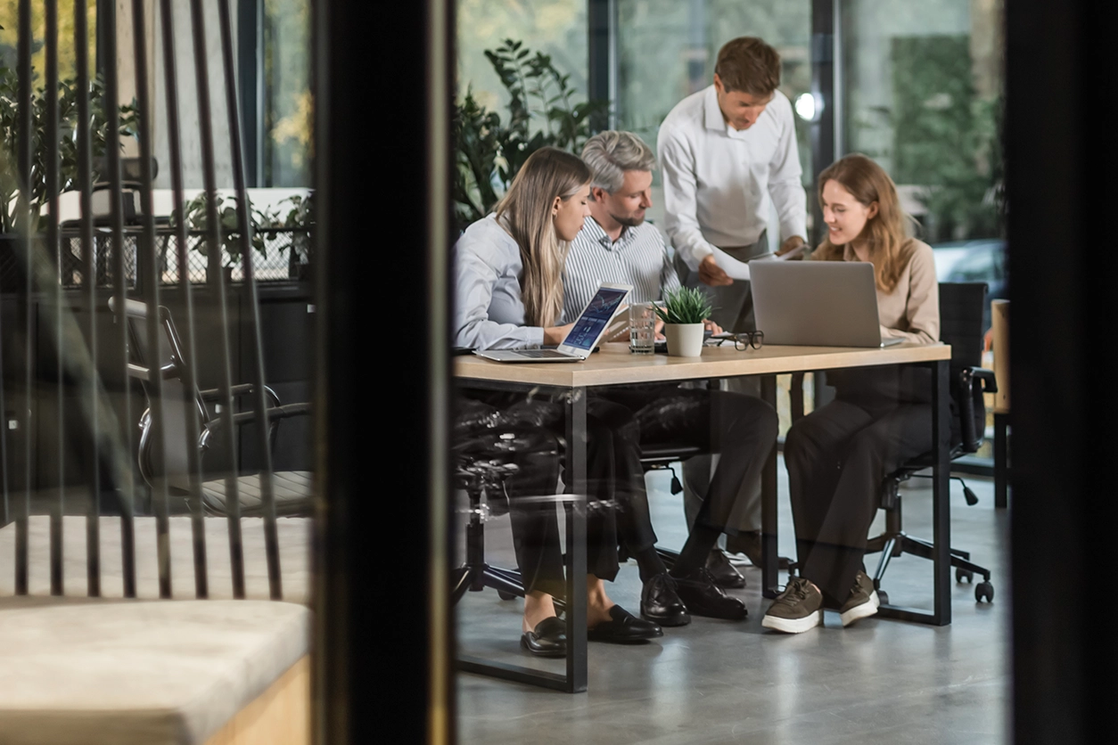 Four people collaborate around a table in a modern office meeting room, reviewing documents and working on laptops, viewed through a glass wall.