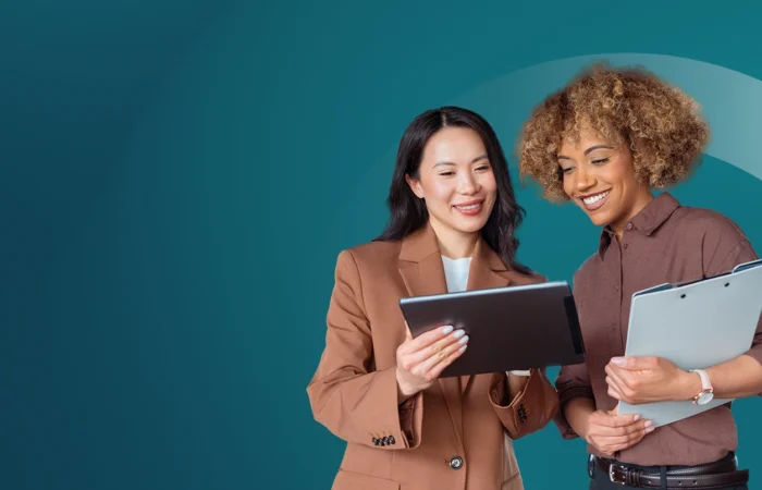 Two professionals standing together reviewing information on a tablet, with one holding a clipboard against a teal background.